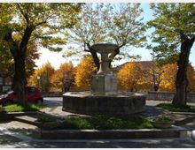 Fontana in pietra con vasca circolare al centro di una piccola piazza pavimentata, circondata da aiuole e alberi. Sullo sfondo si vedono un muretto in pietra e case basse tra vegetazione autunnale.