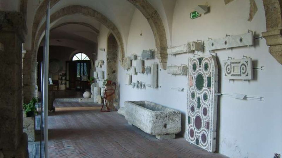 Corridoio interno con archi in pietra e soffitto a volta, pavimento in mattoni e pareti bianche su cui sono esposti frammenti architettonici e pannelli decorativi. Lungo la parete si vedono una vasca in pietra e vari reperti, con una porta ad arco in fondo al passaggio.