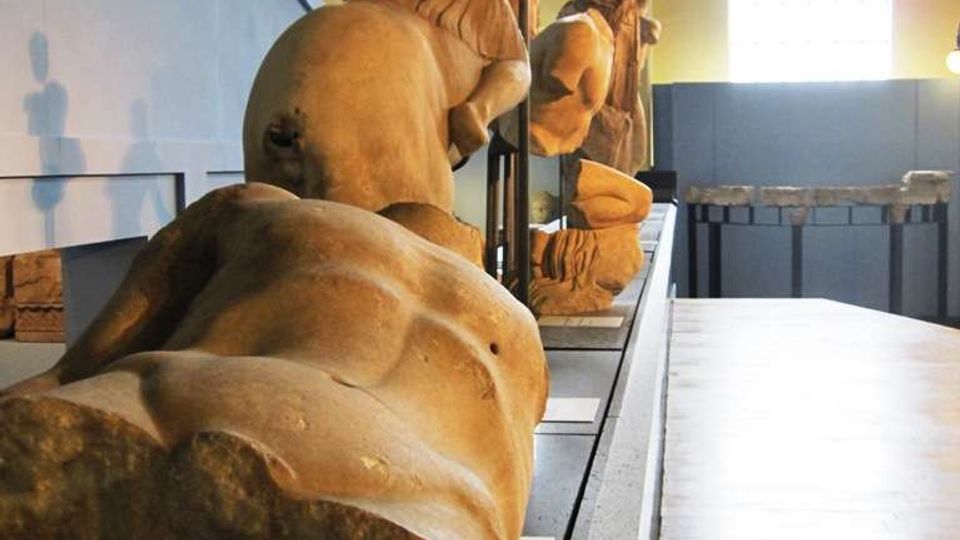 Interno di un museo o galleria con una lunga pedana espositiva che ospita grandi statue in pietra disposte in fila. L’ambiente ha pareti chiare, alti soffitti e ampie finestre che illuminano il corridoio con pavimento in legno.