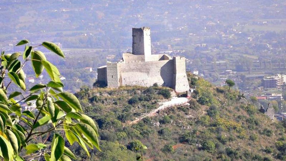 Castello in pietra con torre centrale e mura merlate su una collina boscosa, con sentiero d’accesso visibile. Sullo sfondo si estende una valle con campi e insediamenti urbani leggermente velati dalla foschia.