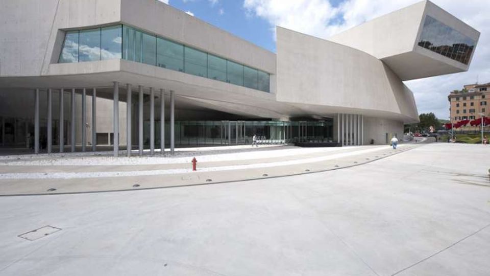 Edificio moderno in cemento chiaro con volumi sporgenti e grandi vetrate orizzontali, sostenuto da una fila di pilastri sottili. Davanti si estende un’ampia piazza pavimentata con gradinate basse e aiuole, sotto un cielo parzialmente nuvoloso.