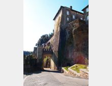 Ingresso ad arco in muratura lungo una strada in salita, ai piedi di una scogliera rocciosa con alti muri di contenimento. Sopra l’arco si vedono edifici storici in pietra con finestre e un camminamento fortificato che segue il bordo della rupe.