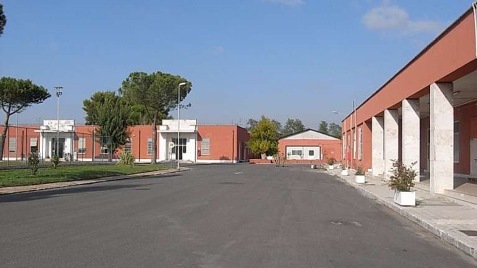 Ampio viale asfaltato in un complesso di edifici bassi in mattoni rossi, con un portico a colonne bianche sul lato destro e piccoli vasi lungo il marciapiede. Sullo sfondo si vedono altri fabbricati, alberi e lampioni sotto un cielo azzurro con poche nuvole.