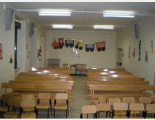 Sala polifunzionale con file di lunghe panche e sedie in legno disposte verso una parete frontale. Soffitto con luci al neon, finestre laterali e decorazioni a tema trenino appese alla parete di fondo.