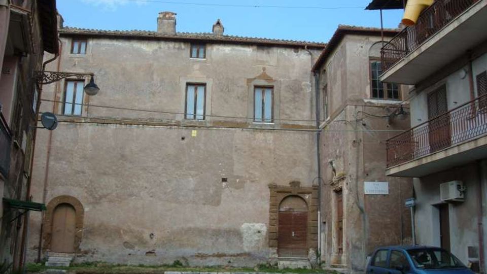 Piazzetta urbana con un grande edificio in muratura intonacata e invecchiata, con finestre strette e portali in pietra, affiancato da palazzi con balconi e ringhiere in metallo. In primo piano si vedono la pavimentazione, un’auto parcheggiata e lampioni a parete sotto un cielo sereno.