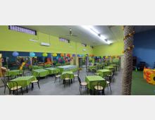 Ampia sala interna per feste per bambini con pareti verde e blu, tavoli coperti da tovaglie verdi e sedie nere, illuminazione a neon e ventilatori a soffitto. Palloncini e decorazioni colorate sono appesi lungo la parete con specchi, con area giochi e strutture per bambini sullo sfondo.