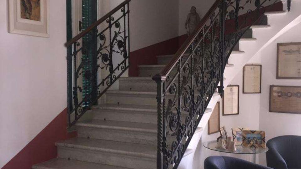 Interno di un edificio con scala in pietra che sale su due rampe, dotata di ringhiera in ferro battuto decorata. Pareti bianche con fascia rossa, quadri e cornici appesi, e un piccolo angolo salotto con poltrone e tavolino accanto alla scala.