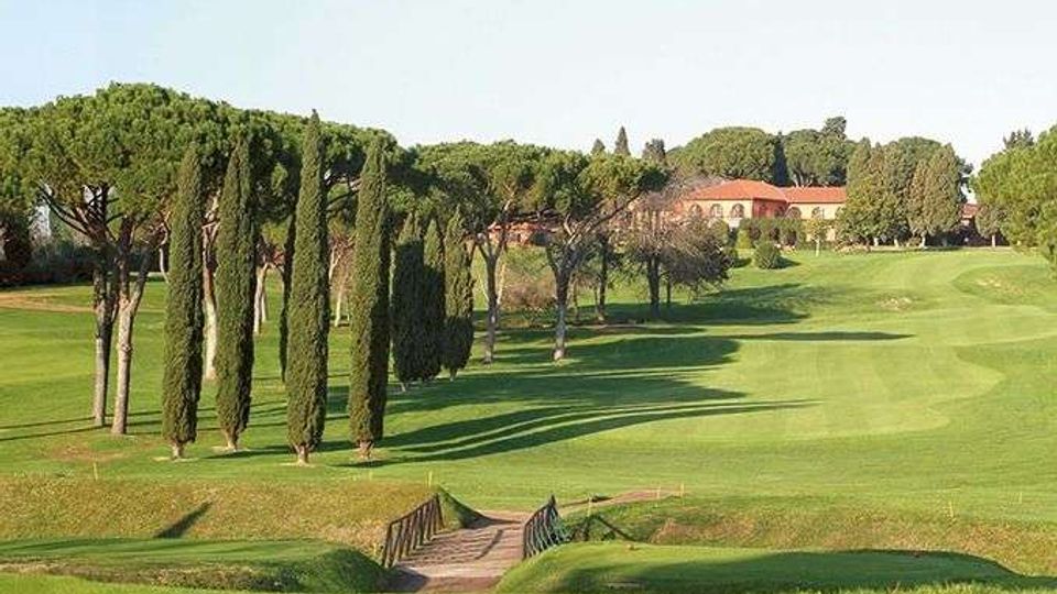Ampio campo da golf con prati ondulati e un piccolo ponte in legno in primo piano. Lungo il percorso si vedono filari di cipressi e pini a ombrello, con edifici rurali dal tetto rosso sullo sfondo.