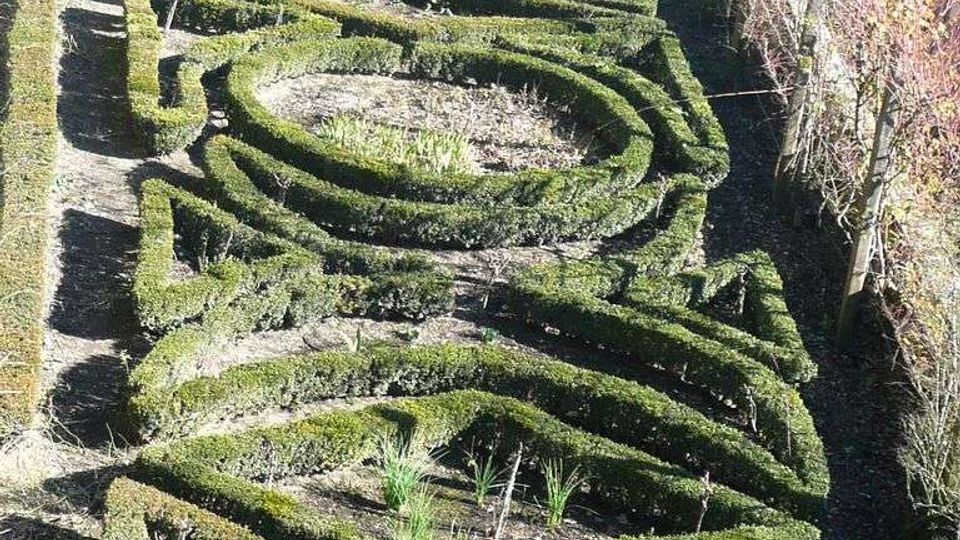 Giardino formale visto dall’alto con siepi di bosso potate a labirinto e aiuole interne, con una piccola fontana e vasca d’acqua nella parte superiore. Il giardino è affiancato da vialetti e confina con un edificio in pietra sul lato destro.