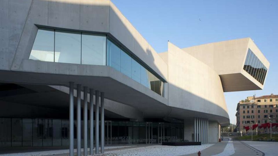 Edificio moderno in cemento e vetro con volumi sporgenti e grandi finestre angolari, sostenuto da sottili pilastri metallici. Davanti si vede una piazza pavimentata con percorso curvo, mentre sullo sfondo compaiono altri palazzi urbani.