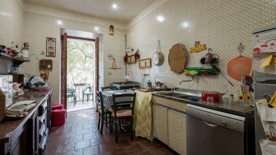 Cucina abitabile con pareti rivestite di piastrelle chiare a motivo geometrico e pavimento in cotto, con tavolo centrale e mobili lungo le pareti. Porta-finestra aperta su un piccolo balcone con sedie e vista su alberi, e zona lavello con lavastoviglie a vista.