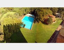 Vista dall’alto di un ampio giardino con grande prato verde e una piscina rettangolare azzurra circondata da alberi. Sul lato destro si vedono edifici con tetto in tegole e portico, e in primo piano un bordo di muratura in mattoni.