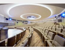 Ampio auditorium moderno con numerose file di poltrone beige disposte a semicerchio attorno a un palco curvo in legno. Il soffitto presenta grandi elementi circolari con illuminazione integrata e faretti, con pareti laterali attrezzate per audio e luci.