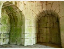 Interno di una struttura in pietra con pareti in blocchi e volte a botte, con due nicchie ad arco incassate e soffitto curvo. La superficie mostra macchie di umidità e patina verdastra, su pavimento in pietra.