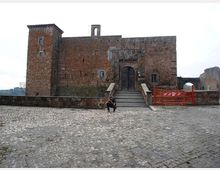 Facciata di un castello in pietra e mattoni con torre laterale e grande portale ad arco, raggiungibile tramite una scalinata con parapetti. In primo piano si vede un ampio piazzale lastricato in pietra, con una zona transennata sul lato destro.