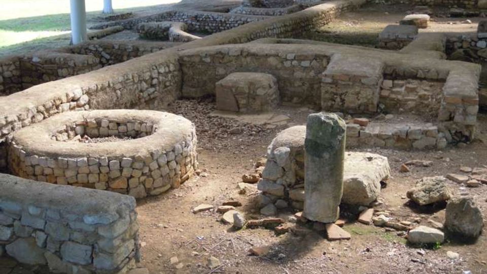 Scavi archeologici all’aperto con muri bassi in pietra che delineano ambienti rettangolari e un’area circolare, sotto una tettoia sostenuta da pilastri. Il terreno è in terra battuta con pietre sparse, e sullo sfondo si vede un’area verde con alberi.