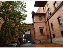 Cortile interno tra edifici in mattoni a più piani, con finestre ad arco e grondaie, affiancato da un grande albero. Un vialetto conduce a un cancello metallico verso la strada, con alcune auto parcheggiate ai lati.
