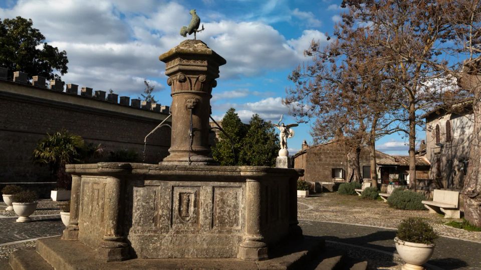 Fontana monumentale in pietra al centro di una piccola piazza lastricata, con colonna decorata e scultura in cima. Intorno si vedono un muro merlato, alberi spogli, panchine e edifici storici sotto un cielo azzurro con nuvole.