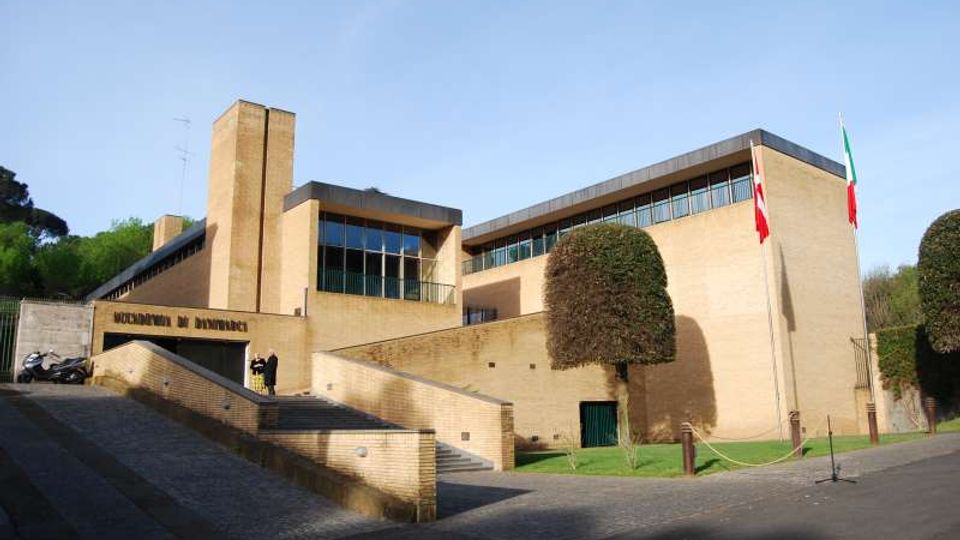 Edificio istituzionale moderno in mattoni chiari con volumi rettangolari, ampie finestre a nastro e una torre verticale; l’ingresso è raggiungibile tramite una rampa e una scalinata. Davanti si trovano un’area pavimentata, prato con alberi potati e aste portabandiera.