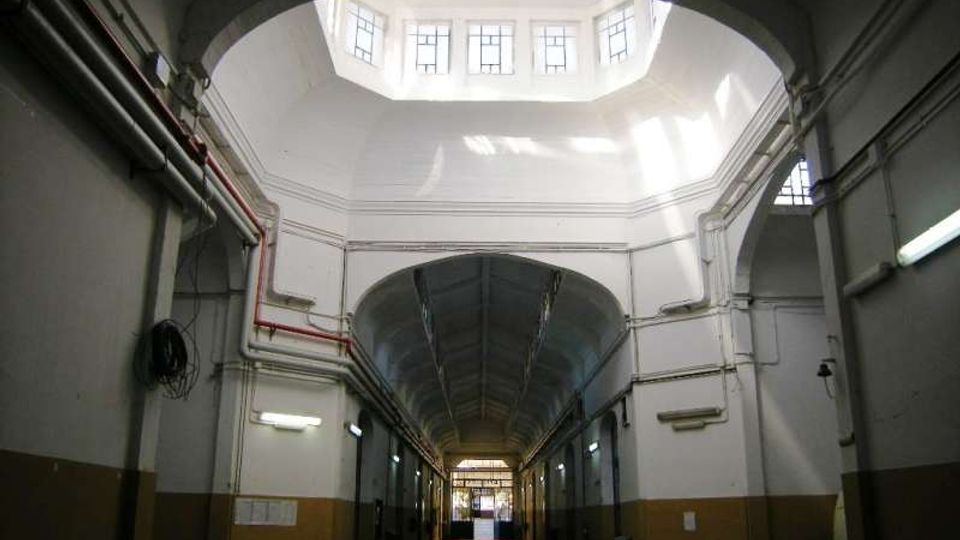 Interno di un lungo corridoio industriale con soffitto alto a volta e grandi archi, pareti chiare con tubazioni a vista e luci al neon. In alto un lucernario con finestre lascia entrare la luce naturale, mentre in fondo si intravede un’uscita con portone.