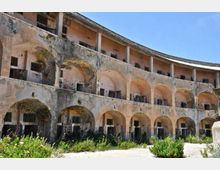 Edificio storico in rovina con facciata curva a più piani, caratterizzata da file di arcate e balconate con intonaco scrostato e finestre sbarrate. In primo piano si vede un cortile con vegetazione incolta e pavimentazione consumata sotto un cielo sereno.