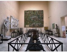 Sala museale con pareti alte color crema, opere d’arte e pannelli espositivi lungo i lati, tra cui grandi rilievi e statue. Al centro si vede un percorso delimitato da ringhiere nere con teche e modellini, e sulla parete di fondo un grande dipinto.