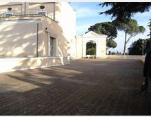 Cortile lastricato con pavimentazione in mattoni a spina di pesce, affiancato da un edificio chiaro con terrazza e finestre. Sullo sfondo si vede un arco monumentale e alberi alti in un’area esterna panoramica.