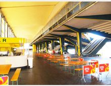 Ampio spazio interno tipo atrio o area ristoro con tavoli e panche in fila, pavimento scuro e chiosco bar laterale. Struttura industriale con colonne e travi in acciaio verniciate, grandi finestre e scale mobili che salgono al piano superiore.