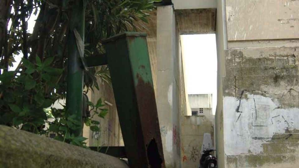 Vicolo stretto tra edifici in cemento con muri scrostati e macchie di umidità, visto dal livello del suolo. In primo piano si vedono vegetazione e un contenitore metallico arrugginito, con pavimentazione in cemento e un’apertura luminosa in fondo.
