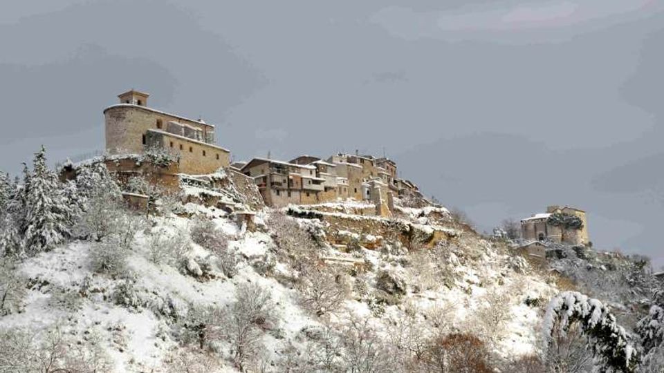 Borgo in pietra arroccato su un crinale montano, con edifici storici e una struttura tipo fortezza o castello. Il pendio sottostante è coperto di neve e alberi innevati, sotto un cielo grigio invernale.