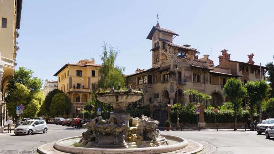 Rotatoria urbana con grande fontana in pietra al centro, circondata da strade con auto parcheggiate e alberi. Sullo sfondo si vede una villa storica in stile liberty con torre, archi e tetti in tegole, affiancata da edifici residenziali.