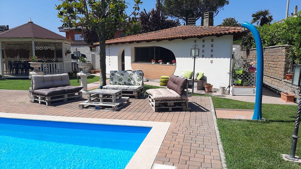 Cortile di una villa con piscina rettangolare azzurra in primo piano e pavimentazione in mattoni. Sullo sfondo ci sono divani da esterno su pallet, un gazebo bianco e un piccolo edificio con tetto in coppi e veranda, circondati da prato, piante e muri di confine.