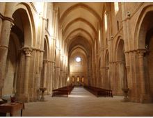 Interno di una chiesa in pietra con lunga navata centrale, arcate su colonne e volte a ogiva. File di panche ai lati conducono verso l’altare, illuminato da una finestra rotonda e da strette finestre verticali sul fondo.