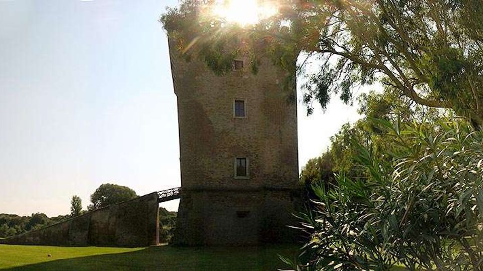 Torre in mattoni di forma quadrata in un’area verde, con un lungo muro in pietra che si estende lateralmente. Alberi e cespugli incorniciano la scena, con il sole che filtra tra le fronde sullo sfondo.