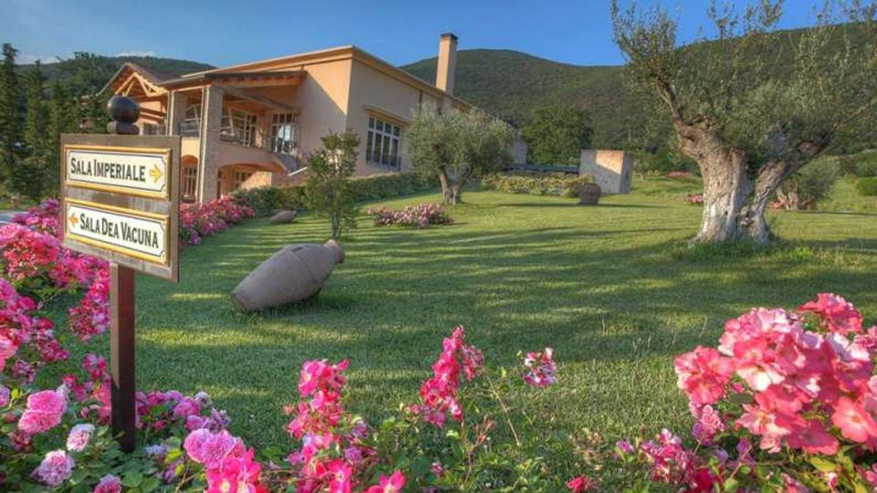 Giardino curato con prato verde e aiuole di fiori rosa, ulivi e un grande anfora decorativa, con un edificio moderno a due piani sullo sfondo e colline boscose dietro. In primo piano un cartello direzionale indica “Sala Imperiale” e “Sala dei Vacuna”.