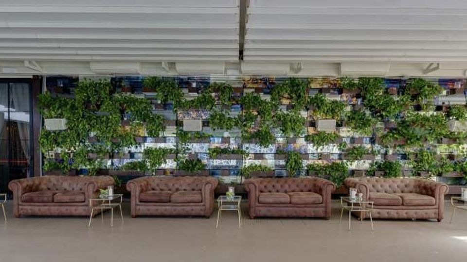 Ampia area lounge interna con soffitto a pannelli bianchi e fila di divani capitonné marroni affiancati da piccoli tavolini in metallo. Sullo sfondo, una grande parete verde con piante e moduli decorativi a griglia.