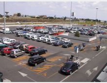Ampio parcheggio asfaltato all’aperto con molte auto parcheggiate in file ordinate, segnaletica orizzontale con frecce direzionali e stalli riservati. Sullo sfondo si vedono strade di scorrimento, lampioni alti e un’area collinare con edifici sparsi.