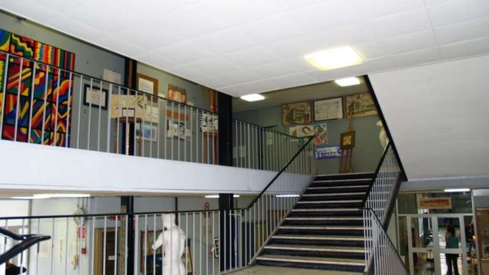 Atrio interno di un edificio pubblico con pavimento in marmo chiaro, ampio vano scala centrale con ringhiere metalliche e un pianerottolo su due livelli. Pareti decorate con pannelli colorati e cornici, illuminazione a plafoniere e porte vetrate sullo sfondo.