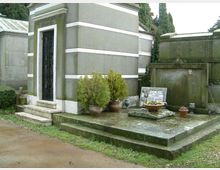 Cappella funeraria in un cimitero, con edificio in muratura grigia a fasce bianche e porta metallica raggiunta da pochi gradini. Davanti c’è una piattaforma in cemento con lapide, vasi di fiori e piccoli arbusti, circondata da altri loculi e cipressi sullo sfondo.