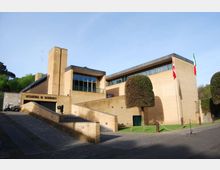 Edificio istituzionale moderno in mattoni chiari con volumi rettangolari, ampie finestre a nastro e una torre verticale; l’ingresso è raggiungibile tramite una rampa e una scalinata. Davanti si trovano un’area pavimentata, prato con alberi potati e aste portabandiera.