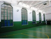 Interno di una palestra con pavimento verde segnato da linee gialle per campi sportivi e pareti bianche con fascia verde. Lungo un lato ci sono alte finestre ad arco con telai verdi e reti appese, sotto un soffitto alto con travi a vista.