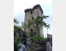 Torre medievale in pietra con merlature sulla sommità e piccole aperture, vista dal basso. In primo piano una scalinata di pietra tra rocce e vegetazione conduce verso la torre, con un lampione e alberi ai lati.