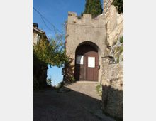 Porta d’ingresso in mattoni di un borgo medievale, con grande arco a tutto sesto e portone in legno lungo una strada acciottolata in salita. Ai lati si vedono mura in pietra, vegetazione e cavi aerei sotto un cielo sereno.