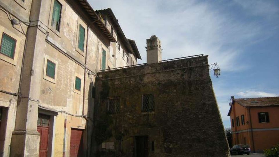 Cortile urbano con edifici in muratura intonacata e pietra: a sinistra una palazzina a più piani con finestre verdi e portoni al piano terra, al centro un edificio più basso in pietra scura con piccolo comignolo. Sullo sfondo si vede una strada con una casa moderna e un lampione sotto un cielo parzialmente nuvoloso.