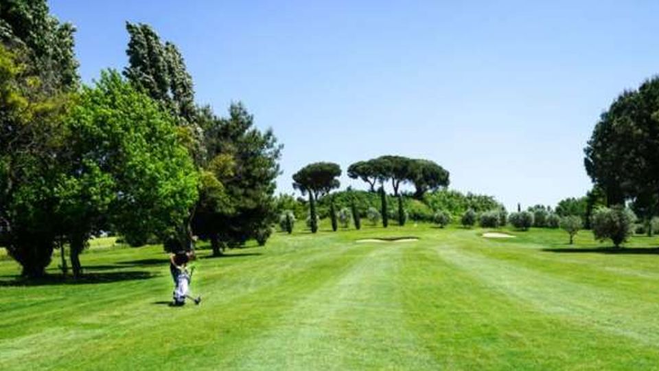 Ampio campo da golf con fairway in erba rasata e bunker di sabbia, circondato da alberi e cespugli. Sullo sfondo si vedono pini a ombrello sotto un cielo azzurro limpido.