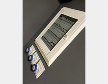 Quadro elettrico da parete con sportello trasparente, contenente interruttori e moduli su guide DIN, montato su una parete grigia. Accanto sono visibili tre prese industriali blu su placche bianche.