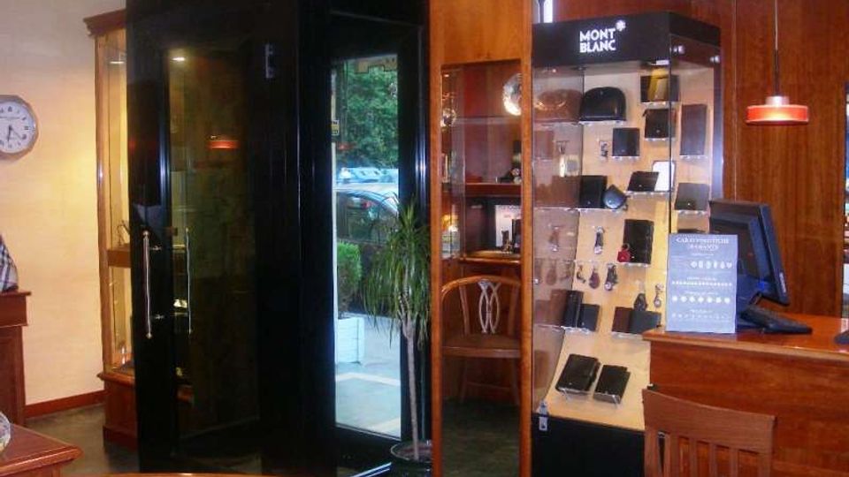 Interno di un negozio con arredi in legno, bancone e vetrine espositive; al centro c’è una cabina in vetro nera e, sulla destra, una teca illuminata con accessori e articoli del marchio Montblanc.