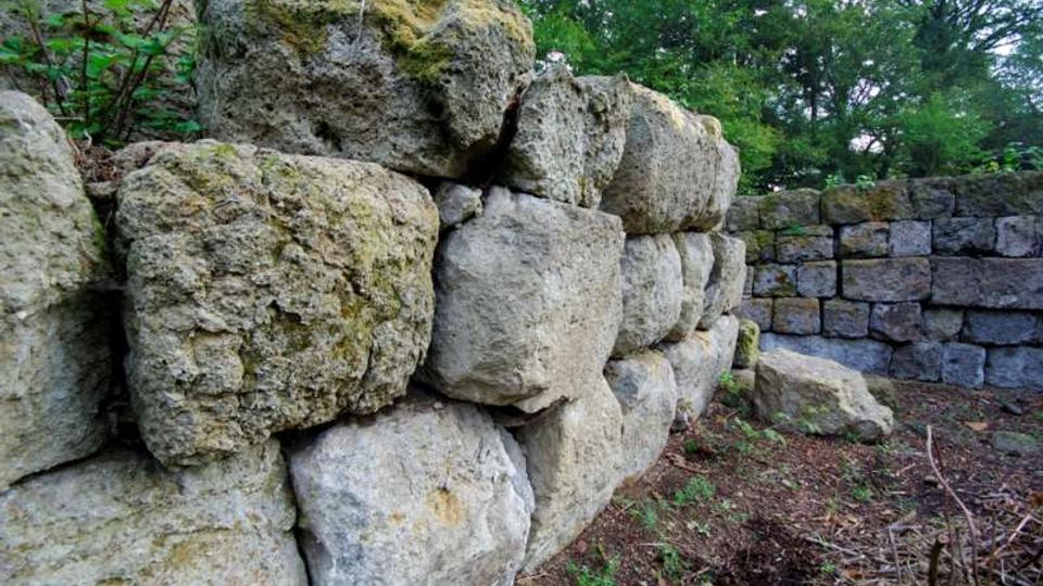 Muro in pietra a secco composto da grandi massi irregolari sovrapposti, con un tratto di muratura in blocchi squadrati sullo sfondo. L’area è all’aperto, su terreno di terra con vegetazione bassa e alberi fitti intorno.
