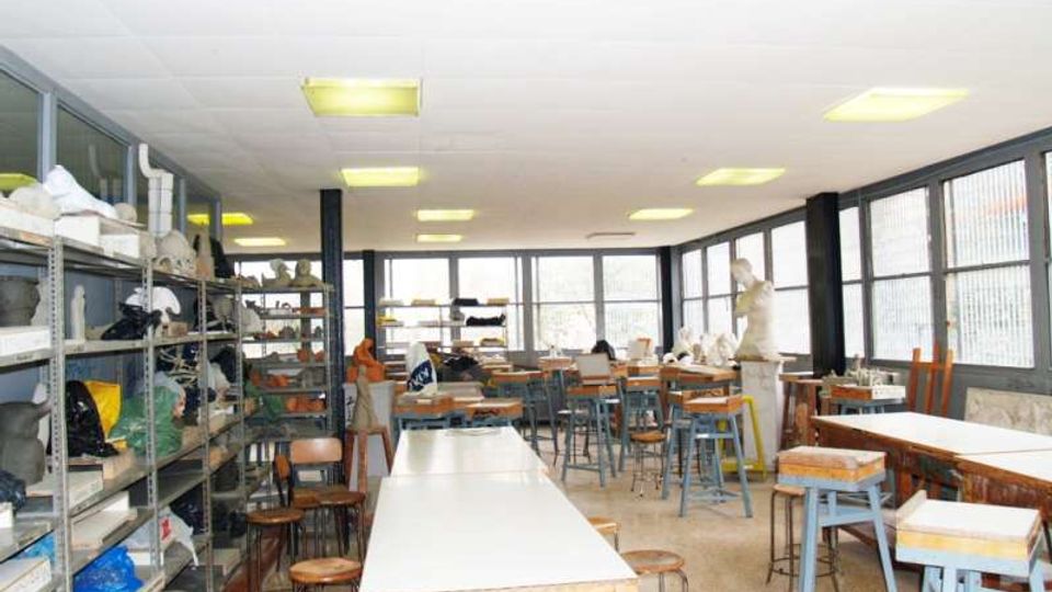 Aula laboratorio artistico con grandi tavoli da lavoro e sgabelli, scaffalature laterali piene di materiali e oggetti. Ampie finestre lungo una parete illuminano l’ambiente, con colonne scure e lampade fluorescenti a soffitto.