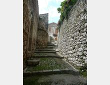 Vicolo in pietra in salita con gradini irregolari e pavimentazione acciottolata, fiancheggiato da alti muri in muratura grezza. In fondo si vede la facciata di una casa con porta ad arco e finestre, con vegetazione che sporge dal muro a destra.
